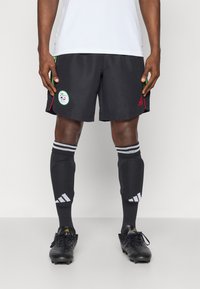 Černé fotbalové šortky s zelenými a červenými akcenty, se stylovým kruhovým logem. Černé ponožky s bílými pruhy Adidas. Černé kopačky.