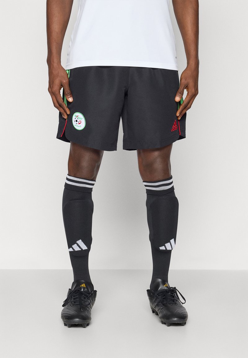 Černé fotbalové šortky s zelenými a červenými akcenty, se stylovým kruhovým logem. Černé ponožky s bílými pruhy Adidas. Černé kopačky.