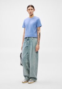 Svetlo modra bombažna majica s kratkimi rokavi, v kombinaciji z ohlapnimi svetlo prani jeans hlačami. Na roki so vidne značilne tetovaže; srebrni čevlji.