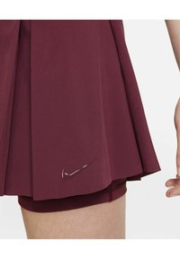 Bordeauxrode geplooide sportrok met elastische tailleband en binnenbroekjes, met een glanzend Nike-logo op de linkeronderkant.