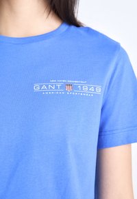 Apykaklė mėlyna medvilninė marškinėliai su baltu logotipu, kuriame yra "GANT" ir šarvo dizainas su raudonais ir mėlynais akcentais.