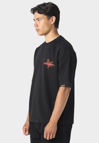 Jonge man in een zwart oversized T-shirt met een rood diagonaal gestreept ontwerp en tekst op de linkerborst, staande tegen een effen achtergrond.