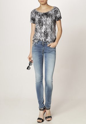 Žena v černo-bílém tričku s potiskem, světle modrých úzkých džínách, černých sandálech na podpatku a s brýlemi na slunce v jedné ruce.