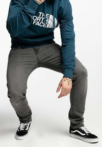 Tmavě modrozelená mikina s bílým logem, šedé slim-fit džíny a černo-bílé tenisky. Výrazné tetování na rukou.