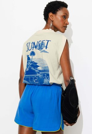 Personne aux cheveux courts portant un débardeur avec "SUNSET" et un motif côtier, un short bleu avec une bordure jaune, tenant un sac noir.