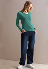 Maglietta a maniche lunghe di colore teal con scollatura a barca, abbinata a jeans a gamba larga blu scuro e sneakers bianche, posata su uno sfondo neutro.