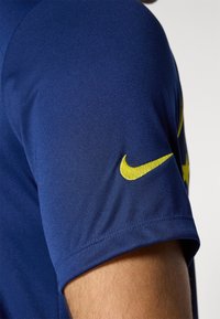 Marineblå atletisk skjorte med korte ærmer, prydet med et gult Nike-logo på højre ærme; glat stofstruktur og tætsiddende design.
