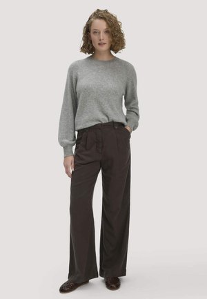 Frau mit lockigem Haar steht vor einem schlichten Hintergrund und trägt einen grauen Langarmpullover, dunkel weite Hose und braune Slip-On-Schuhe.