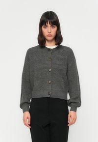Donna con capelli scuri, che indossa un cardigan grigio a bottoni e pantaloni neri, in piedi davanti a uno sfondo bianco semplice.