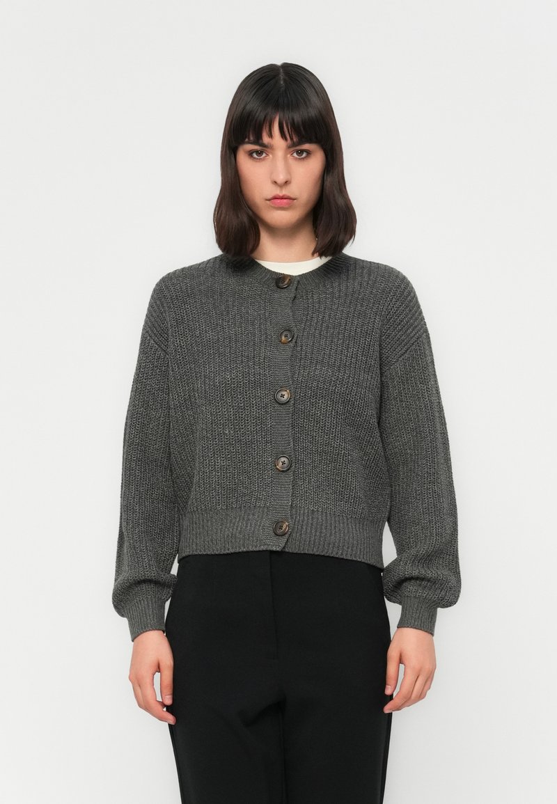 Donna con capelli scuri, che indossa un cardigan grigio a bottoni e pantaloni neri, in piedi davanti a uno sfondo bianco semplice.