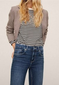 Blazer de patrón de cuadros marrón, camisa de manga larga blanca y negra a rayas, y jeans de denim azul. El conjunto presenta una silueta ajustada.