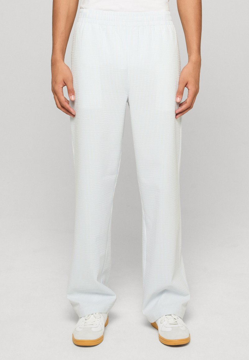 Personne portant un pantalon droit texturé bleu clair et des baskets blanches à semelles en gomme, debout devant un fond uni.
