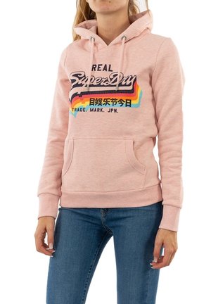 Personne portant un sweat à capuche rose SuperDry avec un motif arc-en-ciel et un texte, associé à un jean bleu, visible de la moitié de la cuisse jusqu'au cou.