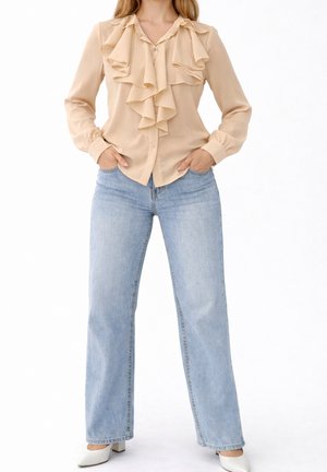 Kobieta nosząca beżową bluzkę z falbankami z długimi rękawami, jasnoniebieskie jeansy o prostych nogawkach, białe buty na obcasie, ręce w kieszeniach.