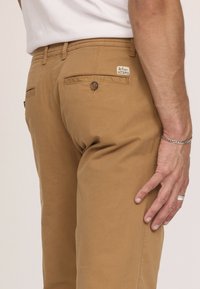 Chinos en coton marron avec une coupe ajustée, dotés d'une poche arrière avec une fermeture à bouton et d'une étiquette de marque près de la taille.