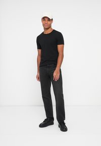 Camisa negra de manga corta combinada con jeans gris oscuro y zapatillas negras. El modelo lleva una gorra de color claro y se encuentra de pie frente a un fondo blanco.