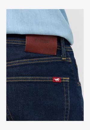 Ciemnoniebieskie dżinsy mają brązową skórzaną metkę z wytłoczonym napisem "MUSTANG" oraz małą czerwoną metkę z białym logo konia.