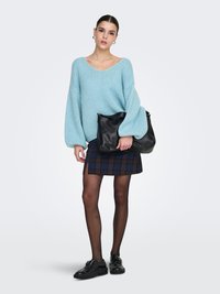 Vaaleansininen neulottu paita, jossa on ilmapallosovitushihat, yhdistetty tummansävyiseen ruudulliseen hameeseen. Musta käsilaukku ja kengät täydentävät asun.