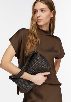 Frau mit langen blonden Haaren, die ein braunes Satin-Oberteil und einen Rock trägt, hält eine große schwarze gewebte Clutch vor weißem Hintergrund.