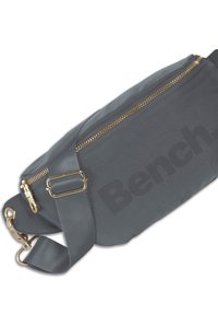 Grijze stoffen schoudertas met een gouden rits, verstelbare schouderband en een opvallend "Bench" logo aan de zijkant. Heeft gouden hardware accenten.