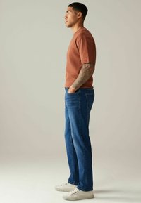 Roestkleurig t-shirt met korte mouwen, gecombineerd met blauwe spijkerbroek en witte sneakers, met een relaxte pasvorm en subtiele stikdetails.