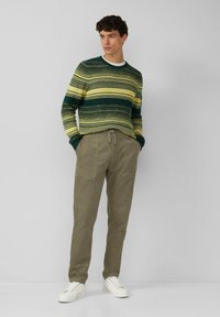 Gebreide trui in groene en gele strepen, met een ronde halslijn; gecombineerd met olijfgroene corduroy broek en witte sneakers.