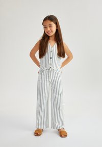 Weiß gestreiftes Outfit mit ärmelloser Bluse und weit geschnittenen Hosen, versehen mit Knöpfen und einem gebundenen Taillengürtel. Getragen mit beigen Slipper-Sandalen.
