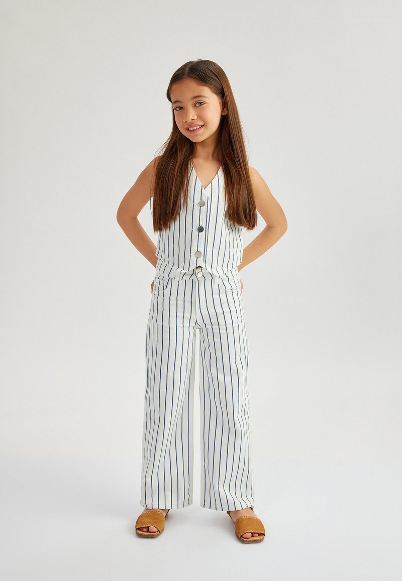 Weiß gestreiftes Outfit mit ärmelloser Bluse und weit geschnittenen Hosen, versehen mit Knöpfen und einem gebundenen Taillengürtel. Getragen mit beigen Slipper-Sandalen.