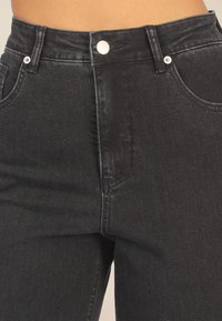Schwarze Jeans aus Denim mit hoher Taille, silbernem Knopfverschluss und zwei Fronttaschen. Die Textur ist glatt mit leichtem Glanz.