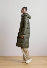 Lang, olivengrøn dunjakke med vandret syninger, hætte og frontlynlås. Sammen med beige bukser og hvide sneakers.