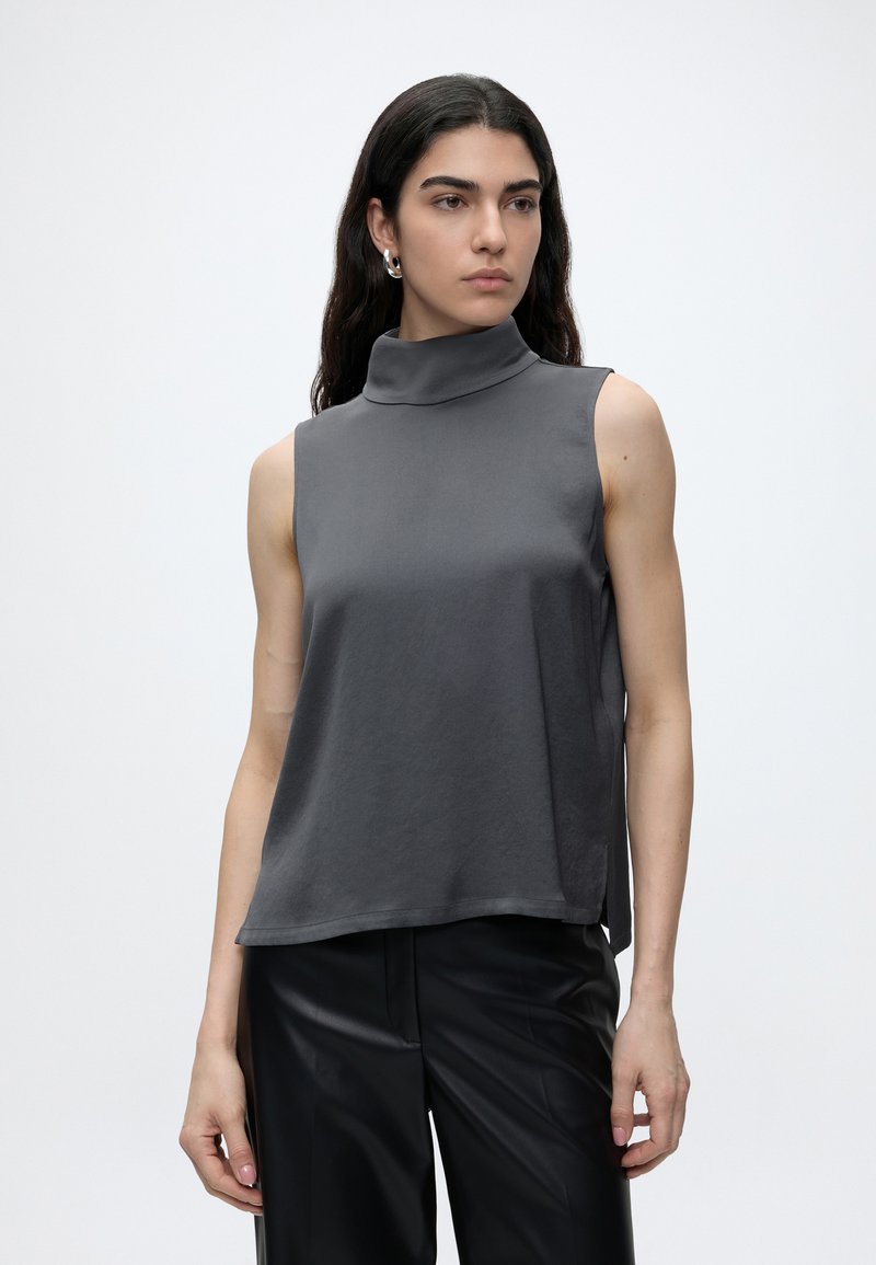 Femme aux longs cheveux foncés portant des boucles d’oreilles argentées en créoles, un haut gris sans manches à col roulé et un pantalon noir sur un fond clair uni.