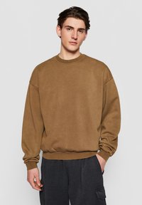 Brun bomulds sweatshirt med rund hals, afslappet pasform, og ribstrikkede kanter og bund. Glat tekstur og minimal detaljering. Kombineret med mørke cargo bukser.