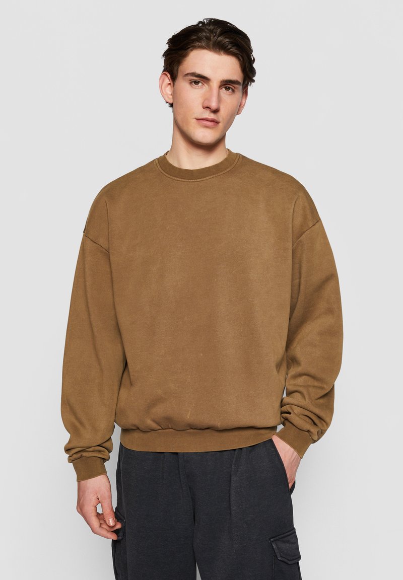 Brun bomulds sweatshirt med rund hals, afslappet pasform, og ribstrikkede kanter og bund. Glat tekstur og minimal detaljering. Kombineret med mørke cargo bukser.
