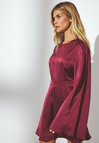 Abito in satin bordeaux con scollatura rotonda e maniche a campana lunghe e fluide; vita aderente e gonna svasata. Texture liscia e lucida.