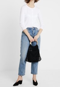 Černá pletená kabelka s dřevěnou rukojetí, nošená s bílou košili na knoflíky a modrými džíny. Černé lodičky s špičatou špičkou doplňují celý outfit.