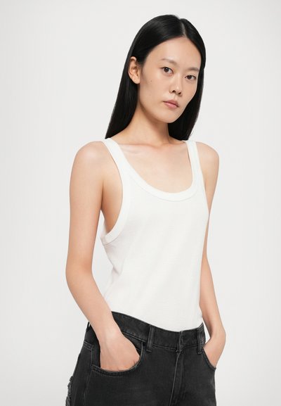 White ribbed tank top with a scoop neckline and wide straps, worn with dark denim jeans. The fabric appears smooth and fitted.