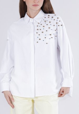 Kobieta ubrana w białą koszulę zapinaną na guziki z długimi rękawami i rozproszonymi metalicznymi koralikami na górnej lewej stronie.