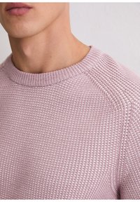 Close-up of a person wearing a light purple textured knit sweater with a crew neckline against a plain background.