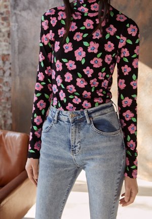 Frau trägt einen schwarzen Rollkragenpullover mit langen Ärmeln und pinkem Blumenmuster sowie hellblaue hoch taillierte Jeans und steht drinnen.