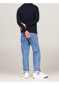 Marineblauwe lange mouw shirt, lichtblauwe baggy jeans en witte sneakers met marineblauwe accenten. Ontwerpelementen zijn onder andere opgerolde jeans en logodetails op de jeans.