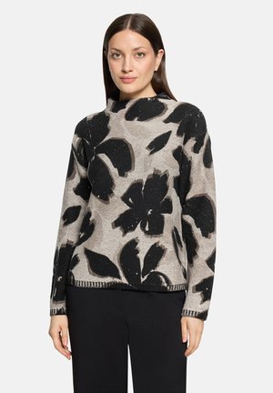 Femme aux cheveux raides et foncés portant un pull beige avec de grands motifs floraux abstraits noirs et un pantalon noir.