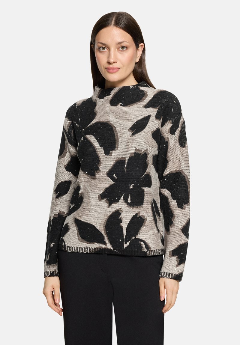 Femme aux cheveux raides et foncés portant un pull beige avec de grands motifs floraux abstraits noirs et un pantalon noir.