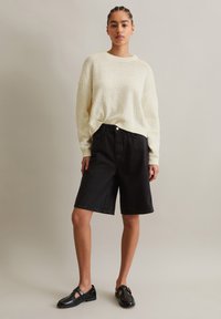 Dan aus cremefarbenem, strukturiertem Strick, schwarzen Denim-Shorts und schwarzen Lederschuhen. Der Pullover hat einen runden Ausschnitt; die Shorts sind knielang und mit einem Knopf ausgestattet.