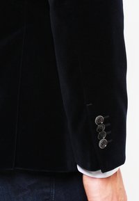 Svart sammet kavajsleeve med sex metallknappar, skräddarsydd passform och kontrasterande vit skjortmudd synlig nedanför. Slät textur.