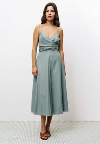 Une robe sans manches, arrivant au genou, en teal atténué, avec un design drapé plissé à la taille, présentant une texture subtile et une silhouette fluide.