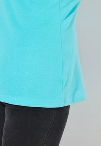 Ein türkisblaues Baumwoll-T-Shirt mit Rundhalsausschnitt, kurzen Ärmeln und geradem Saum, kombiniert mit schwarzen Jeans. Die Textur wirkt weich.