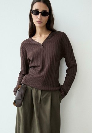 Donna che indossa un maglione a coste color marrone scuro con scollo a V, pantaloni a vita alta verde oliva, occhiali da sole neri slim e una pochette in pelle marrone agganciata ai pantaloni.