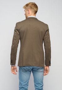 Brun blazer lavet af tekstureret stof, med en tætsiddende design, lange ærmer og sorte knapper på manchetterne, båret over lyseblå jeans.