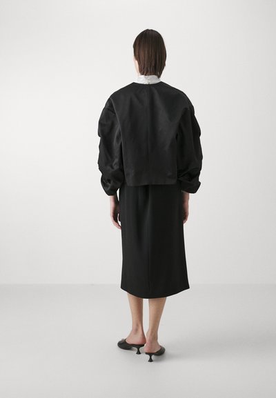 Black cropped jacket with puffed sleeves, paired with a black midi dress and pointy, heel-less shoes, set against a neutral background.