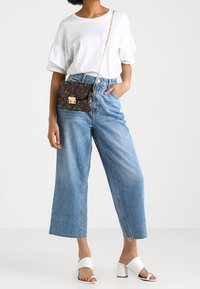 Blommönstrad handväska i mörkröd och grön, med ett guldspänne, bärs över kroppen; kombinerad med en vit volangblus, ljus denimbyxor med vida ben och vita blockklacksandaler.